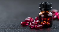 Red omega-3 fish oil capsules spilled from dark amber glass bottle on black stone background, soft focus with copy space for wellness and supplement concepts.