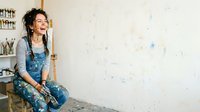 Young woman with dark curly hair and olive complexion, wearing denim overalls and work gloves, sitting against white wall during home renovation project.