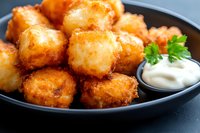 Crispy potato croquettes served on black plate with creamy dip sauce and fresh parsley garnish, golden brown appetizer against dark background.