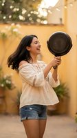Happy young Hispanic woman holding cast iron skillet outdoors in warm sunlight, laughing joyfully while cooking in backyard kitchen space.