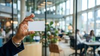 Businessman holding blank white card in modern office lobby with colleagues and glass windows in background, suitable for corporate branding and business communication concepts.