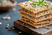 Crispy whole grain crackers with sesame seeds stacked on wooden board, garnished with fresh rosemary sprig. Healthy snack food photography on dark background.