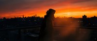 Young man silhouette standing on rooftop overlooking city skyline during dramatic orange sunset. Urban lifestyle and contemplation concept for social media.