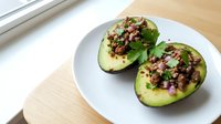 Halved avocado topped with seasoned ground meat, diced red onion, and fresh cilantro on white plate for healthy lunch or dinner meal preparation.