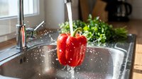 Red bell pepper being rinsed under running water in kitchen sink with fresh herbs and window in background. Cooking preparation and healthy eating concept.
