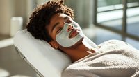 Young African American woman relaxing during spa facial treatment with white sheet mask, lying on massage table in sunlit room, enjoying skincare routine.