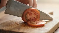 Ripe tomato sliced with sharp knife on wooden cutting board, fresh produce preparation for cooking and meal planning.