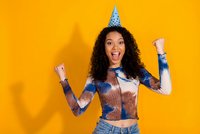 Excited young African American woman with curly hair celebrating birthday party wearing cone hat raising fists in triumph on bright yellow background.