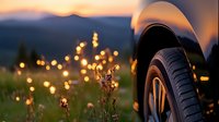 Car wheel against sunset meadow with golden wildflowers and bokeh lights creating dreamy atmosphere. Mountain silhouettes and warm evening glow in background.