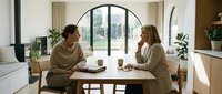 Two women having a meaningful conversation at a wooden dining table in a bright, modern home interior with arched windows and natural light.