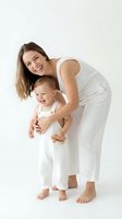 Joyful young Caucasian mother laughing with her happy toddler baby in matching white outfits on a bright white studio background.