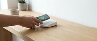 Person with dark skin tone using contactless payment terminal on wooden desk with potted plant in background, representing modern digital transaction technology.
