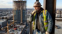 Construction worker in hard hat and safety vest overlooking urban development site from high-rise building during golden hour sunset cityscape view.