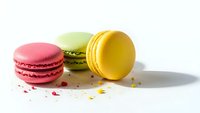 Colorful French macarons in pink, green and yellow on white background with crumbs scattered around for bakery and dessert concepts.