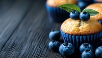Fresh blueberry muffin in blue paper cup decorated with ripe berries and green leaves on dark wooden background, selective focus with copy space.