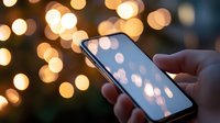 Smartphone displaying glowing bokeh lights on screen, mirroring background holiday illumination. Hand holding device against blurred warm light circles.