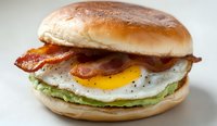 Breakfast sandwich with crispy bacon, sunny side up egg and mashed avocado on toasted brioche bun against light background, close up view of morning meal.