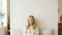 Blonde woman in cream knit sweater sitting on white bed against neutral wall, suitable for lifestyle, wellness, and home decor design projects.