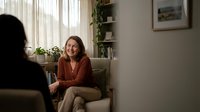 Middle-aged Caucasian woman in rust cardigan sitting comfortably on beige sofa during therapy session in cozy living room with plants and natural light.