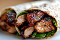 Grilled chicken wrap with fresh spinach leaves and glazed meat pieces in tortilla, close-up view showing juicy texture and caramelized coating.