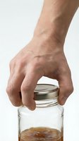 Male hand opening glass jar with metal lid containing honey or jam on white background for kitchen and food preparation concepts.