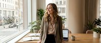Woman in business casual attire standing at wooden desk near window with plants and laptop, suitable for corporate, professional, and workplace concepts.