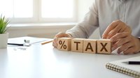Accountant or tax professional arranging wooden blocks spelling tax on white desk with calculator, notebook, and plant, representing tax planning, financial management, and accounting services.