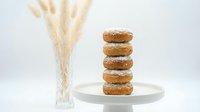 Stack of five golden brown donuts with sugar coating on white plate beside dried wheat stalks against clean white background for bakery and food concepts.