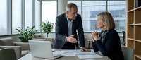 Mature male executive in dark business suit and middle-aged female professional in blazer discussing work at desk with laptop in modern office with city views and wooden shelving.