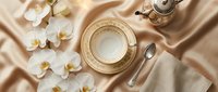 Elegant tea cup with gold trim on satin fabric, white orchids, silver teapot and spoon, luxury table setting for wedding or event.