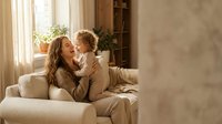 Happy young Caucasian mother and toddler son bonding on beige sofa in bright living room with natural sunlight streaming through window curtains.