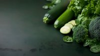 Fresh green vegetables including broccoli, cucumber, and leafy greens arranged on dark background with copy space. Clean eating and healthy food concept.