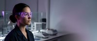 Female scientist wearing protective eyewear in modern laboratory with equipment and purple lighting for research and development work.