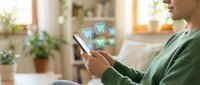 Young woman using smartphone for online shopping with digital cart icons floating above device in cozy home interior with plants and warm lighting.