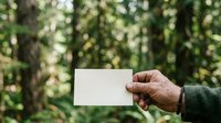 Male hand holding blank white card in forest setting with green trees and natural sunlight, suitable for business messaging and outdoor branding.