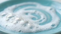 Creamy white foam swirls in turquoise bowl with bubbles creating spiral pattern for skincare cosmetics spa wellness beauty treatment product photography.