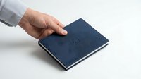 Navy blue hardcover notebook held by a Caucasian male hand on a white surface, showcasing elegant texture for stationery and office supply concepts.