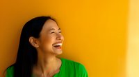 Young Asian woman in bright green top laughing joyfully against vibrant yellow wall background, expressing genuine happiness and positive emotions.