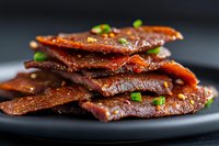 Spicy glazed beef jerky strips garnished with green onions on dark plate, protein snack food for healthy eating and meal preparation.