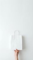 White paper shopping bag held by female hand against minimal light gray background for retail branding and eco-friendly packaging concepts.
