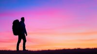 Silhouette of lone hiker with backpack standing on hilltop against vibrant pink and orange sunset sky, dramatic dusk landscape with grass horizon.