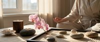 Woman meditating in lotus pose near zen garden with pink orchid, stones, and candles in warm sunlit room, promoting mindfulness and relaxation.
