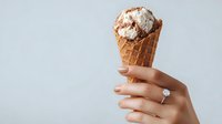Chocolate ice cream scoop in waffle cone held by female hand with engagement ring against neutral gray background for food advertising.