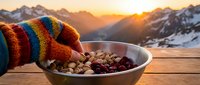 Person wearing colorful striped knit gloves holding bowl of mixed nuts and dried fruits with snow-covered mountains and golden sunrise in background. Outdoor winter snacking.