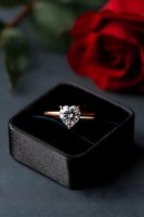 Heart-shaped diamond solitaire engagement ring in rose gold setting displayed in black velvet box with red rose in background, romantic proposal setup.
