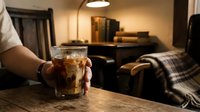 Person holding glass of cola with ice on wooden table in modern living room interior with warm lighting and furniture.
