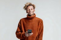 Young African American man in orange turtleneck sweater holding smartphone, smiling and looking away on gray background for lifestyle content.