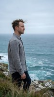 Young Caucasian man in casual gray shirt standing on rocky coastal cliff overlooking ocean waves, contemplative lifestyle portrait for travel and wellness content.