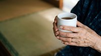 Senior woman with wrinkled hands holding warm beverage cup, wearing dark cardigan. Elderly female enjoying hot drink indoors, relaxation and comfort concept for lifestyle and wellness.