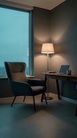Modern home office workspace with dark armchair, desk, laptop, and floor lamp beside large window with blue evening sky view.